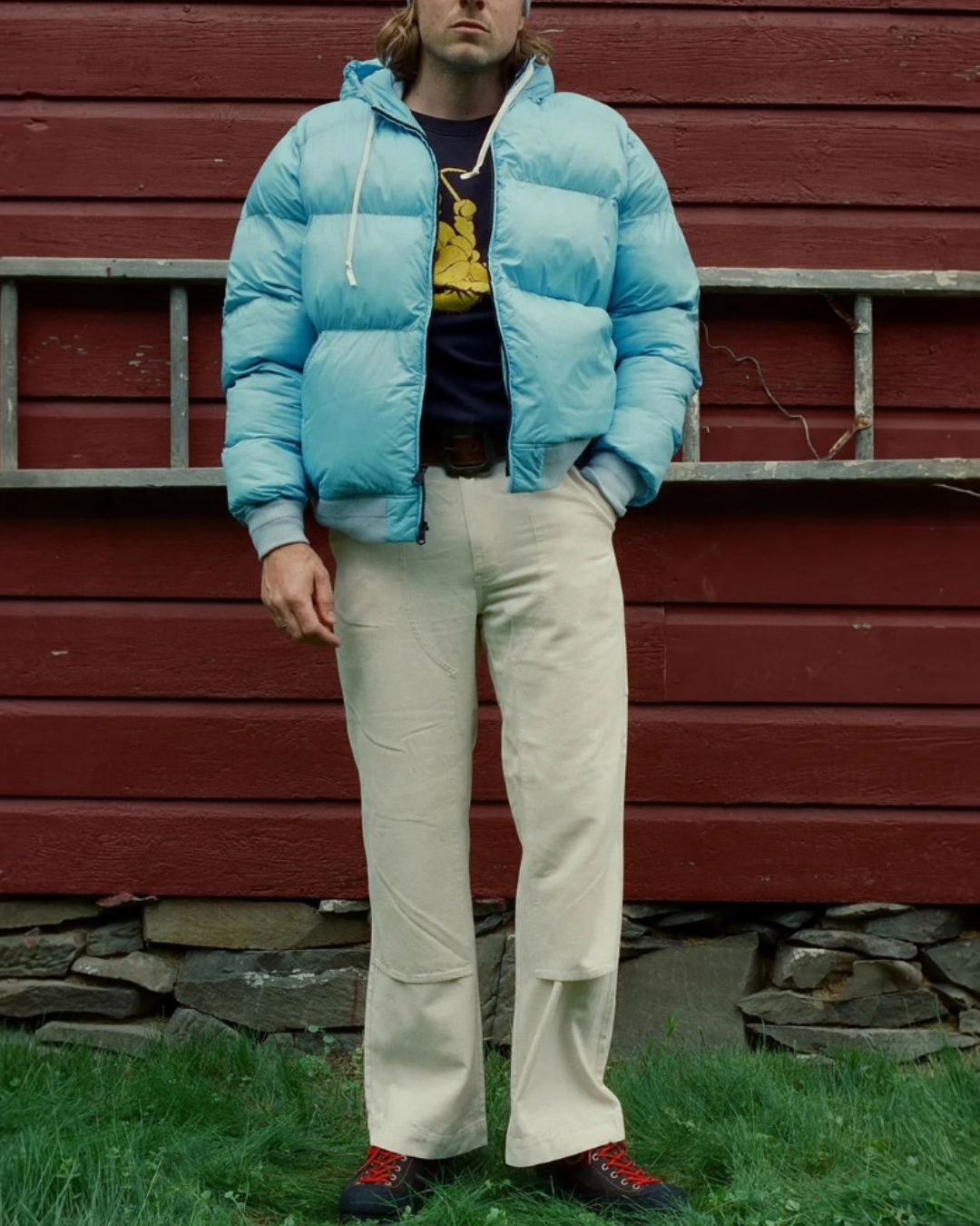 Homme portant la doudoune Alpine Glacier Freeze de Merci Paris, veste en nylon bleu ciel avec capuche devant un mur rouge