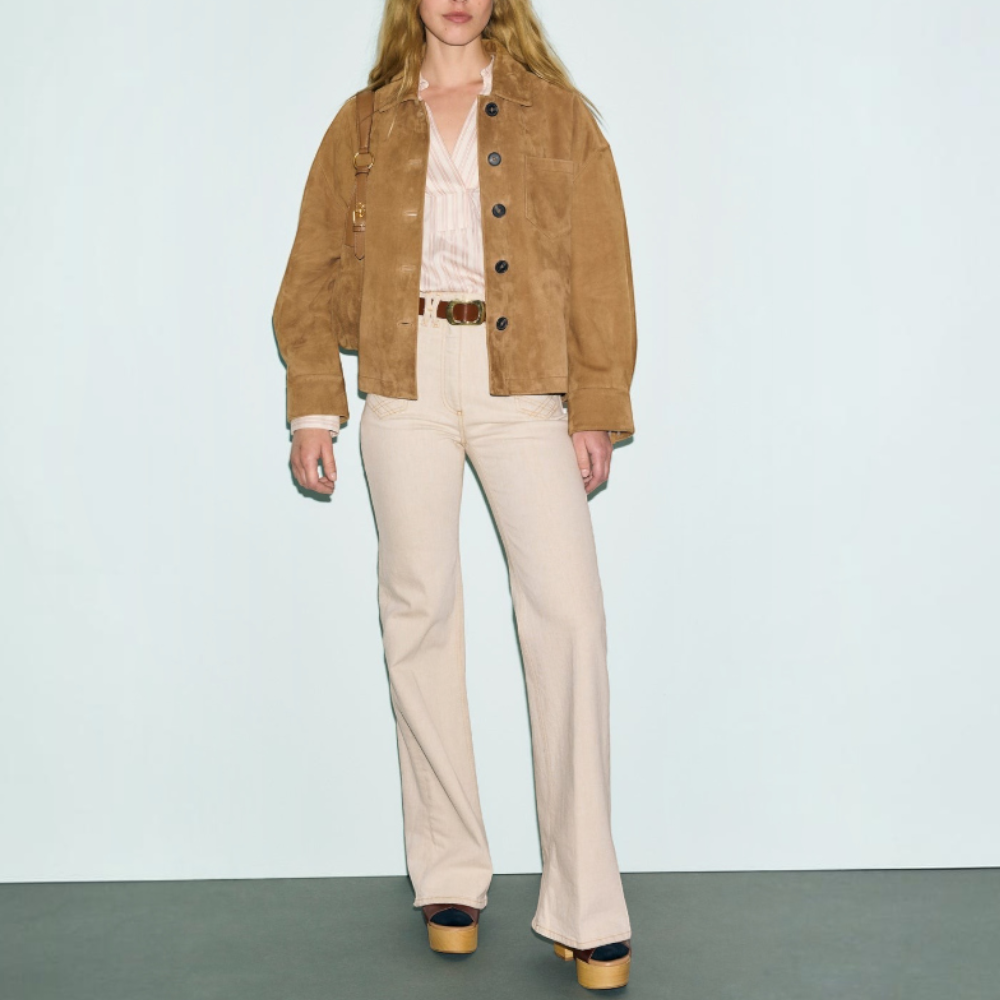 Woman with long blonde hair wears a veste en cuir velours camel, light striped blouse, cream flared pants, a brown belt, and platform shoes. She stands against a plain background, looking at the camera.