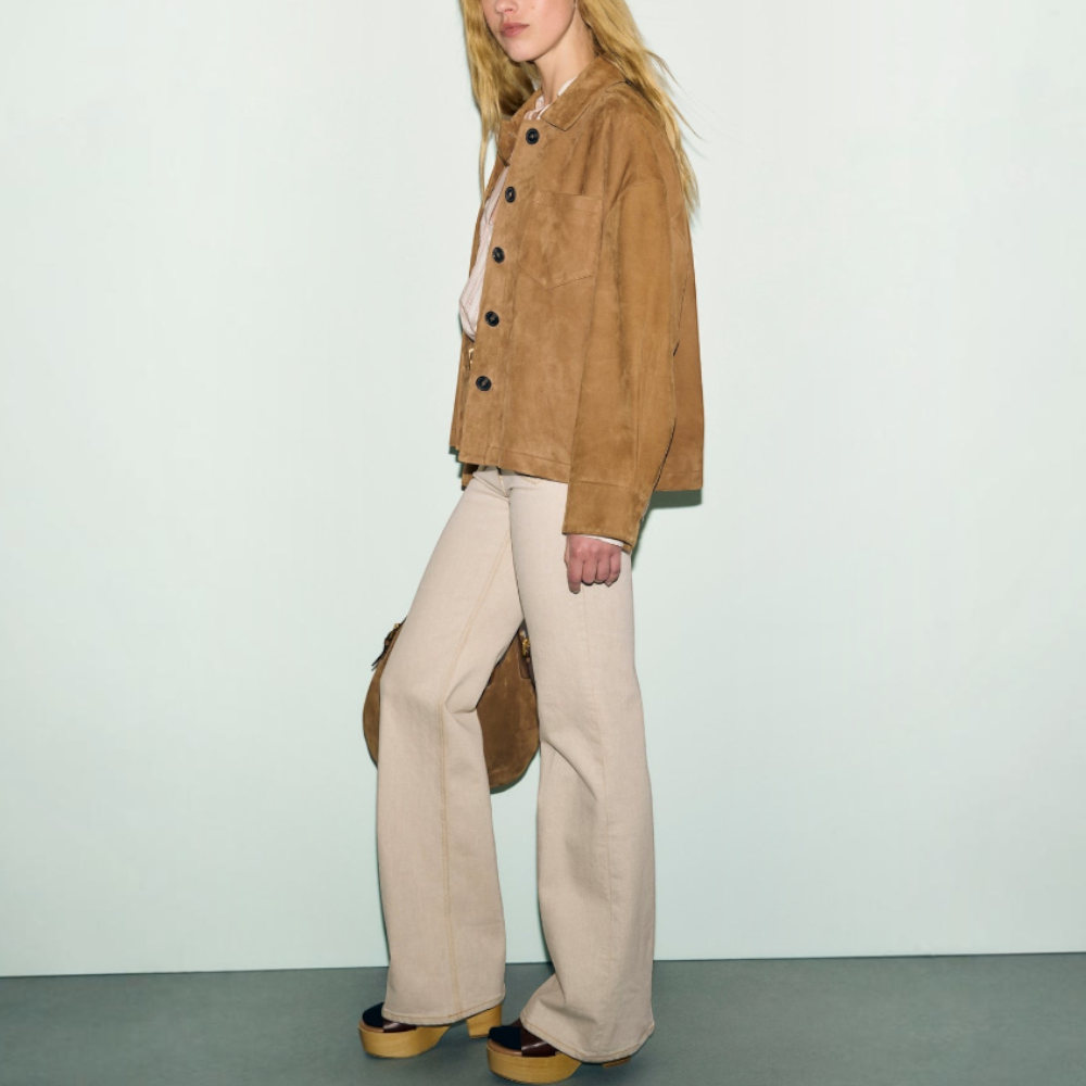 A woman with long blonde hair wears a veste en cuir velours, cream wide-leg pants, and platform shoes, standing against a pale background and holding a matching tan bag.