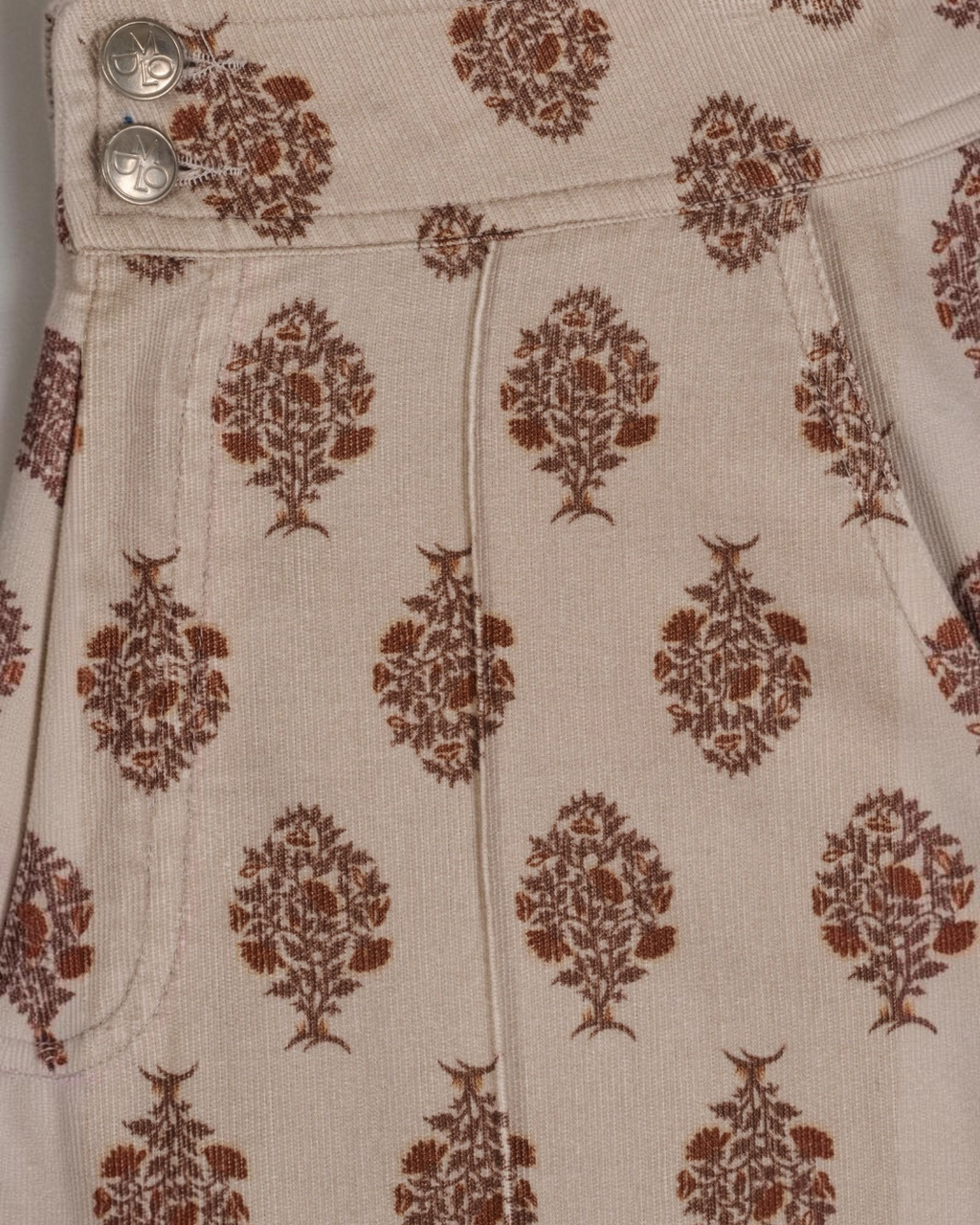 Gros plan sur le pantalon Douglas beige de Merci Paris avec un motif floral marron et des boutons métalliques gravés.