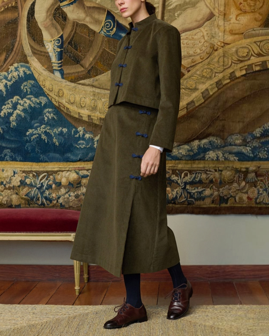 Modèle portant la veste courte kaki Maria De La Orden avec fermetures ornementales devant une tapisserie ancienne