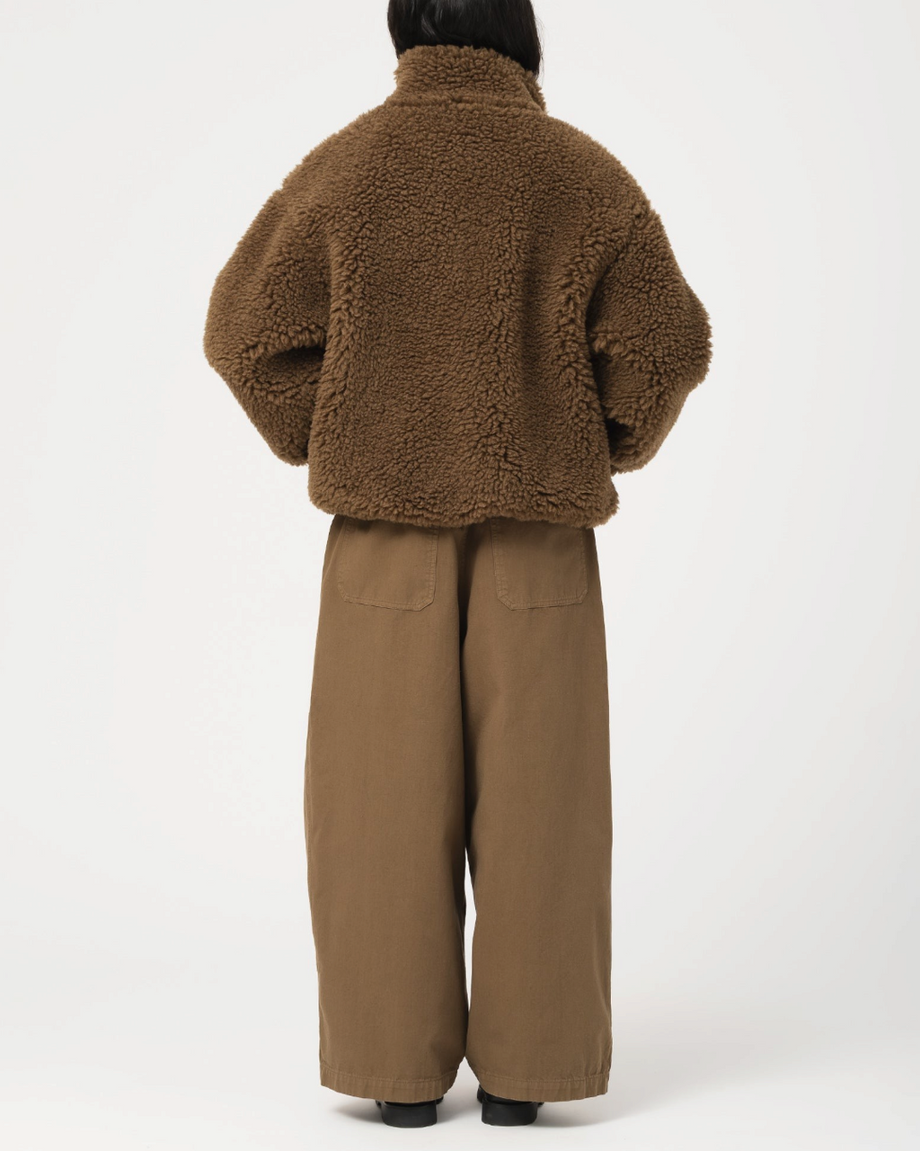Vue arrière d'une femme portant la veste Alpine Jacket en polaire marron Shitake de Merci Paris avec un pantalon large