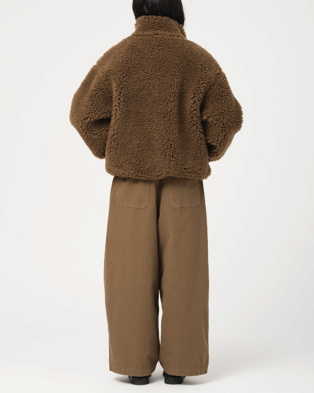 Vue arrière d'une femme portant la veste Alpine Jacket en polaire marron Shitake de Merci Paris avec un pantalon large